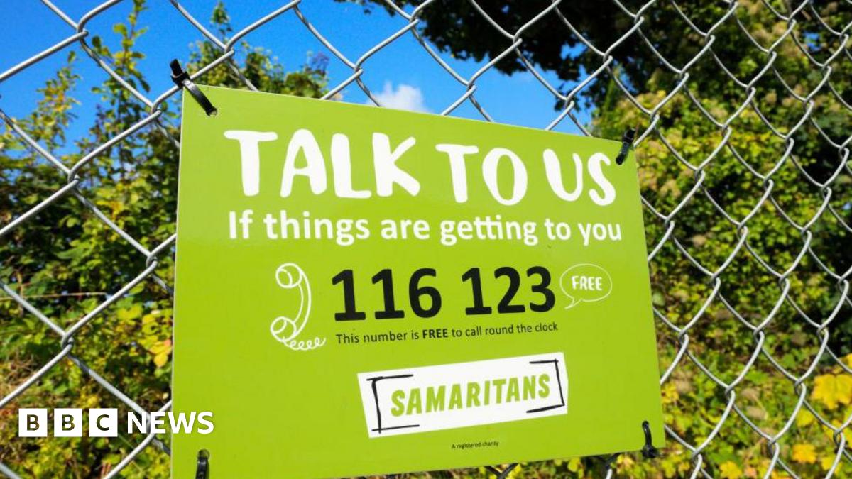 Samaritans sign on a railing in front of some bushes. The sign is green with black writing and gives their contact number.
