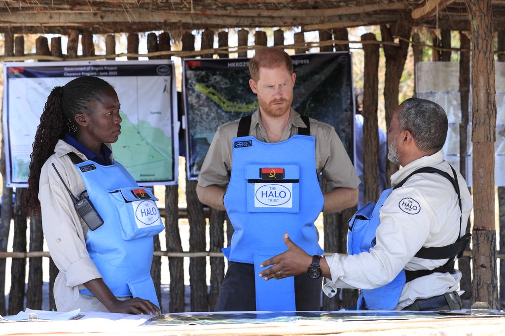 Prince Harry being given a safety talk - he wears a blue safety vest and stands with a woman and a man