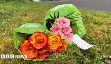 Pink and orange roses wrapped in a green plastic sleeves lie on the grass on a roadside with a white lined piece of paper which says  "We are heartbroken.  May they rest in peace. You are in our prayers."