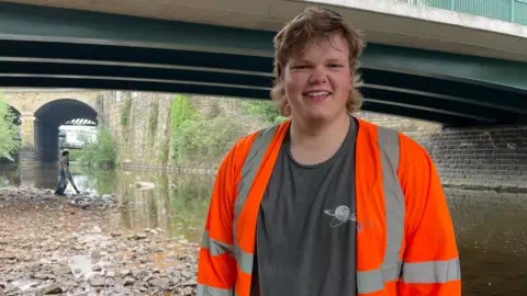 Sheffield City Council A young man with long blonde hair, wearing a grey T-shirt with a hi-vis jacket, smiles for the camera while standing on a river bank.