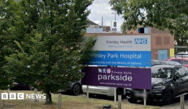 The entrance to Frimley Park Hospital, showing the sign in the car park, with several vehicles parked behind it.