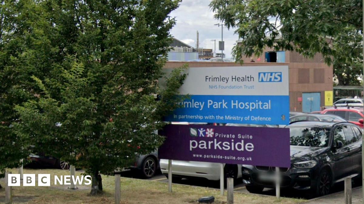 The entrance to Frimley Park Hospital, showing the sign in the car park, with several vehicles parked behind it.