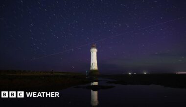 Dark blue night sky showing green and purple colours of the Aurora Borealis visible as a diagonal line of Perseid meteor passed over a white lighthouse