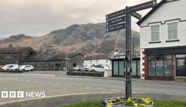A general view of the village of Coniston. There is a sign in the foreground signposting to various attractions in the village. There are shops and buildings lining the main street and the fells in the background.