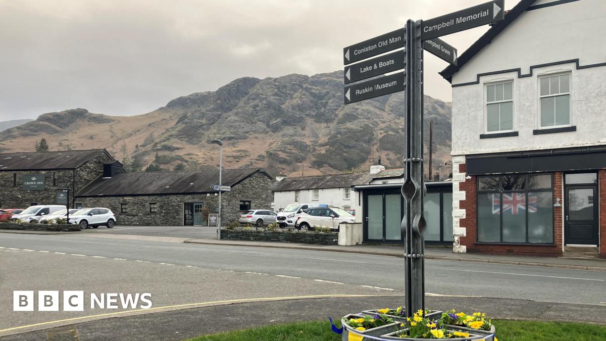 A general view of the village of Coniston. There is a sign in the foreground signposting to various attractions in the village. There are shops and buildings lining the main street and the fells in the background.