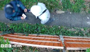 A still of CCTV footage which looks down on two figures - one of whom appears to be holding bank notes. They stand on a path surrounded by shrubbery with a brown fence on one side. The individual on the left is blurred and you cannot see either individual's face.