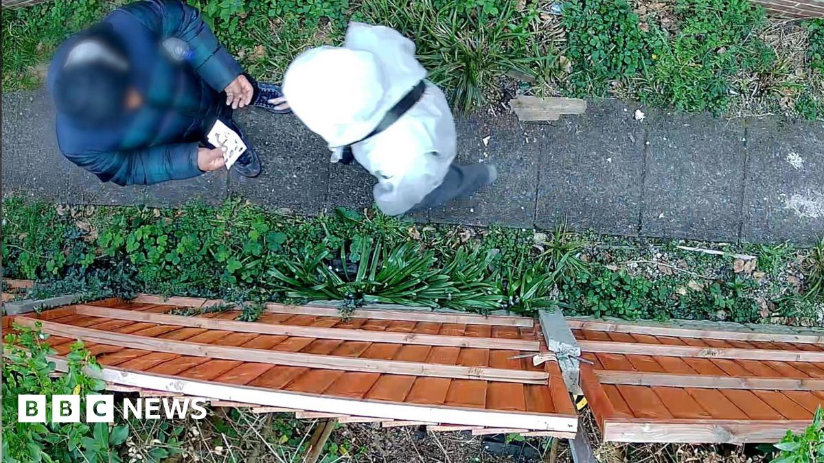 A still of CCTV footage which looks down on two figures - one of whom appears to be holding bank notes. They stand on a path surrounded by shrubbery with a brown fence on one side. The individual on the left is blurred and you cannot see either individual's face.