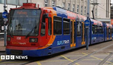A blue and red tram. The doors are closed and the electronic sign at the front reads, 'Halfway'.