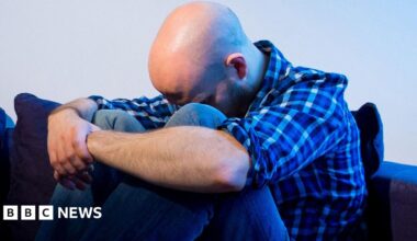 Bald man in a blue shirt sitting on a sofa with his arms around his legs and head down.