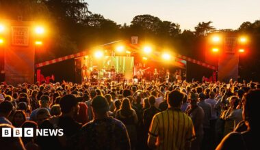 Crowds gather at a stage with artists playing. Signs on either side of the stage say "Mostly Jazz, Funk and Soul Festival". Orange lights are flashing as the crowd plays
