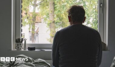 A silhouette of a man sitting on his bed, looking out of the window. He is looking away from the camera. A plant is on the windowsill.