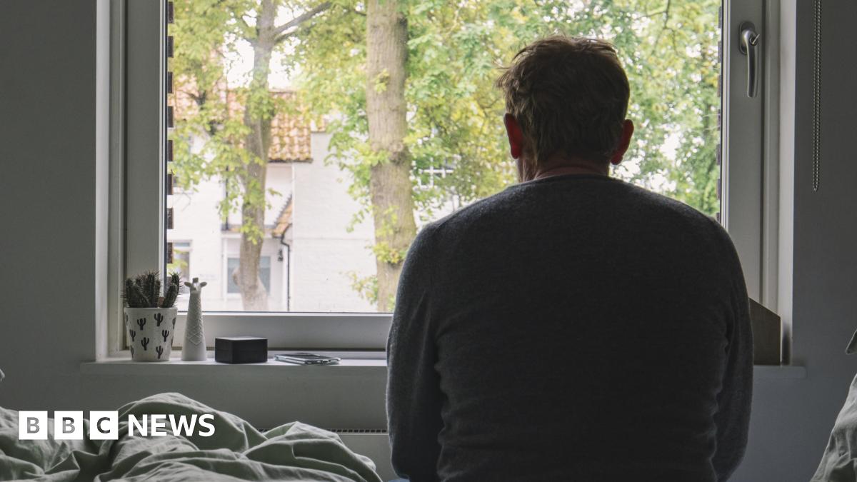 A silhouette of a man sitting on his bed, looking out of the window. He is looking away from the camera. A plant is on the windowsill.