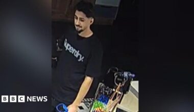 A low quality image of a man wearing a black t-shirt with the words "Superdry" across his chest in white or silver lettering. He has black hair, and a beard across his upper lip and chin. He appears to be carrying some form of bag.