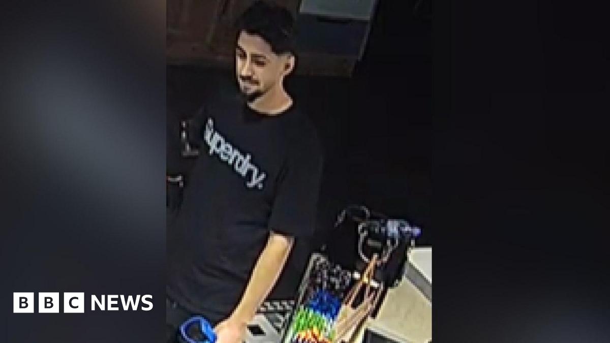 A low quality image of a man wearing a black t-shirt with the words "Superdry" across his chest in white or silver lettering. He has black hair, and a beard across his upper lip and chin. He appears to be carrying some form of bag.
