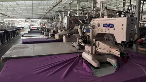 Ed Habershon / BBC An industrial sewing machine sits on purple cloth. In the backgrounds dozens of other similar machines can be seen in a warehouse -  no-one is working at the machines.