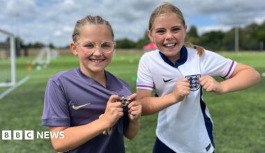 Two young girls, both of them wearing England football tops, one grey and one white. They are both holding the three lions crest on their tops as they smile for the camera.