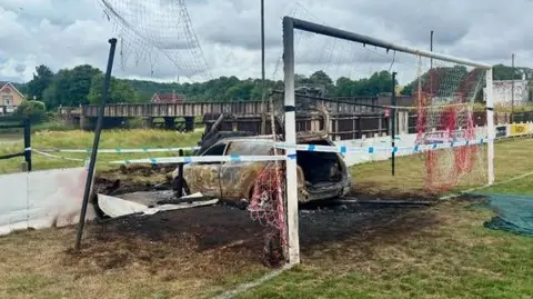 Bristol Manor Farm FC An image of a white car that has been completely burnt out, it is sitting inside a goal post on a football pitch. The goal post and its net are badly damaged, and a patch of grass around the vehicle scorched. The hoarding around the pitch is also damaged where the car has driven into it. 