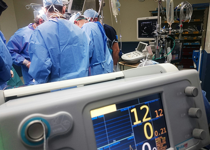 Medical team performing surgery in an operating room with vital signs monitor showing patient’s heart rate and status.