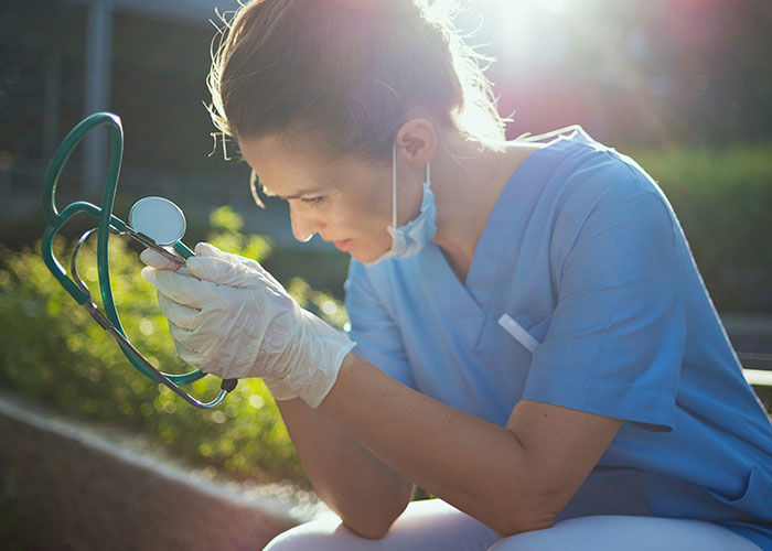 Female healthcare worker in scrubs and gloves looking stressed while holding a stethoscope outdoors, highlighting delayed patient help risks.
