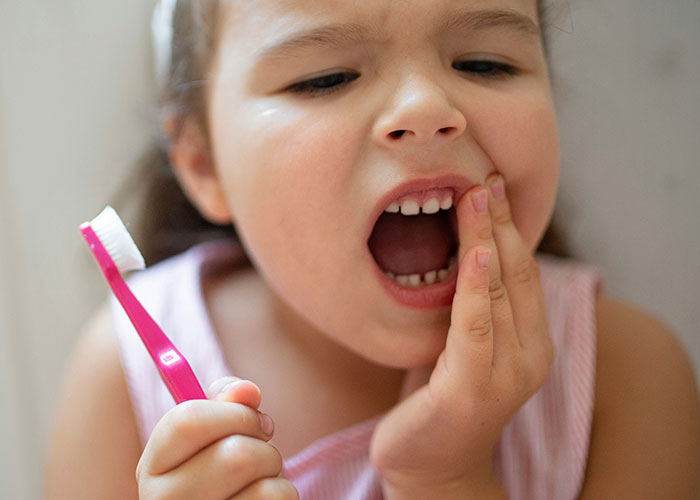 Young girl holding a toothbrush and touching her cheek, illustrating patients delaying help with shocking consequences.