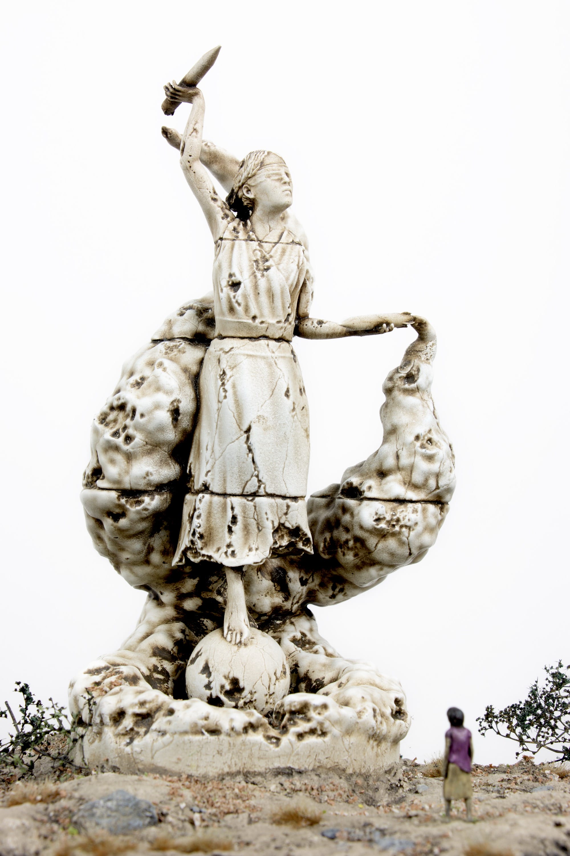 a miniature person sitting at the base of a figurative statue who is blindfolded with a spear in her hand and an explosion coming from her hand