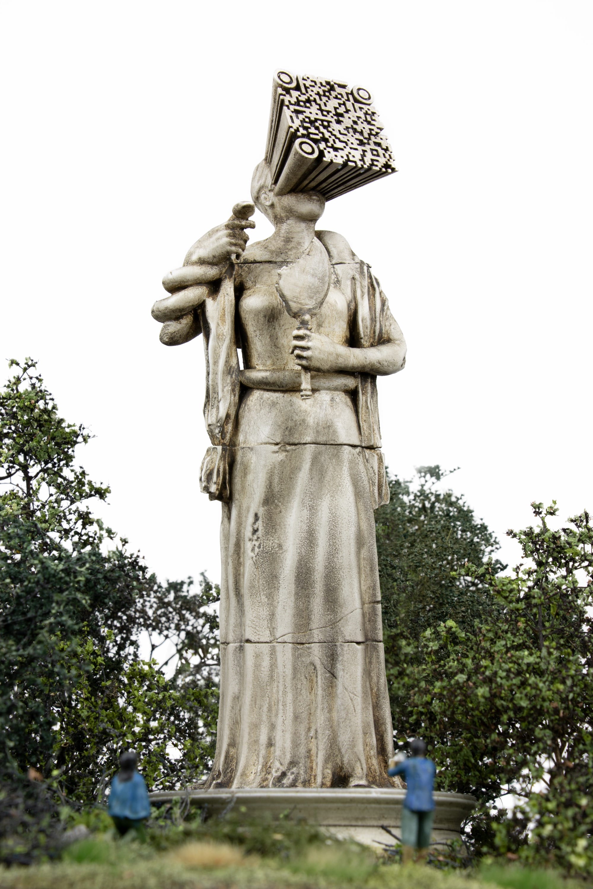 a miniature person sitting at the base of a figurative statue whose face is a QR code