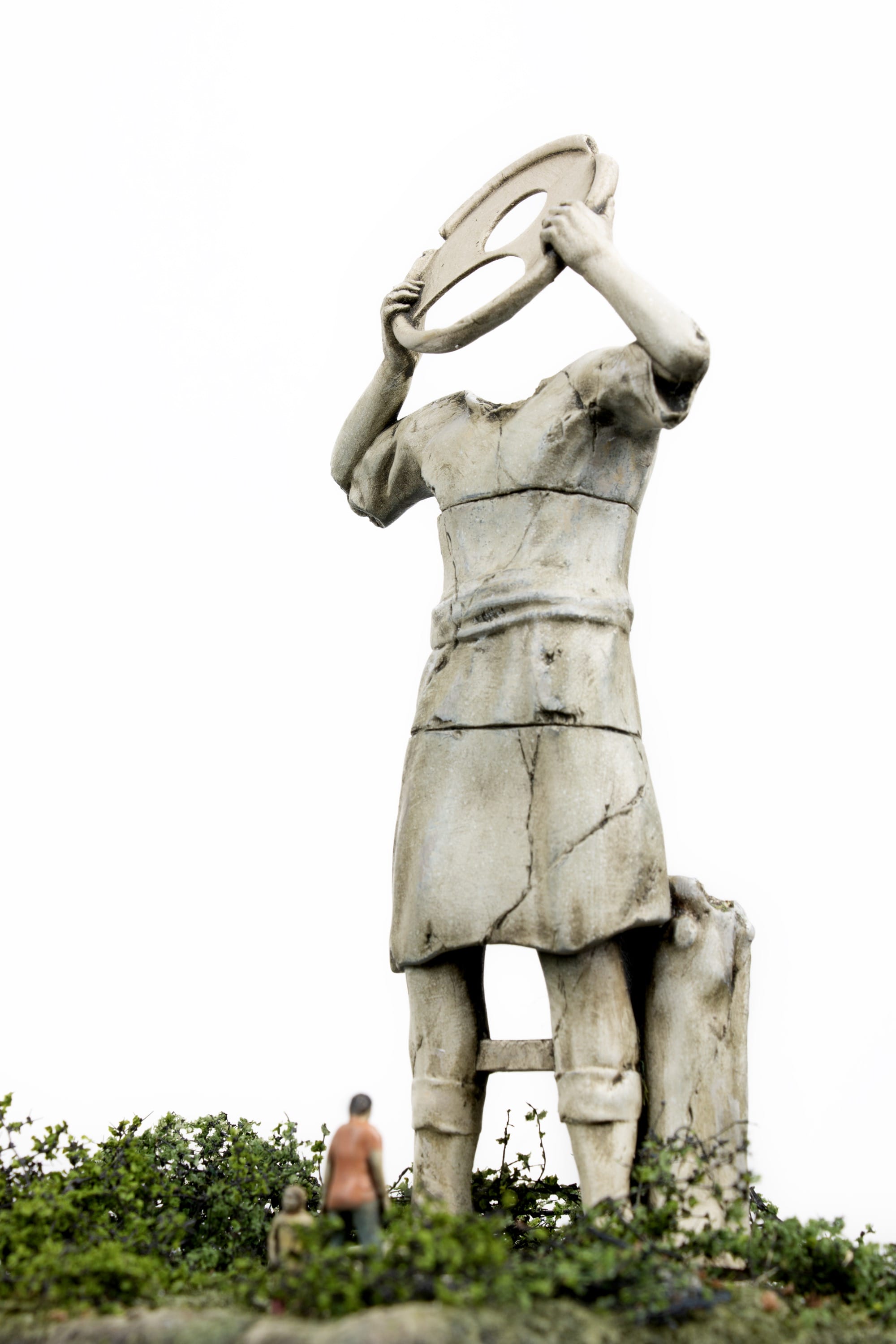 miniature figures look up at a headless antique statue holding a round object