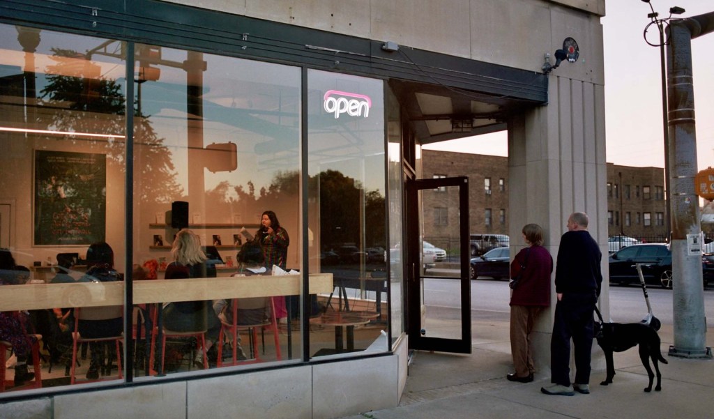 Two people stand with a dog outside the entrance to Dream Palace Books. Through large glass windows, we see seated people listening to someone read from a book on stage.