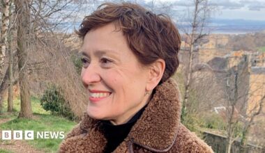 Carys Davies is smiling as she looks to the left of the camera. She has short brown hair and is wearing a black top with a brown fleece jacket. She is in the countryside, with trees behind her.