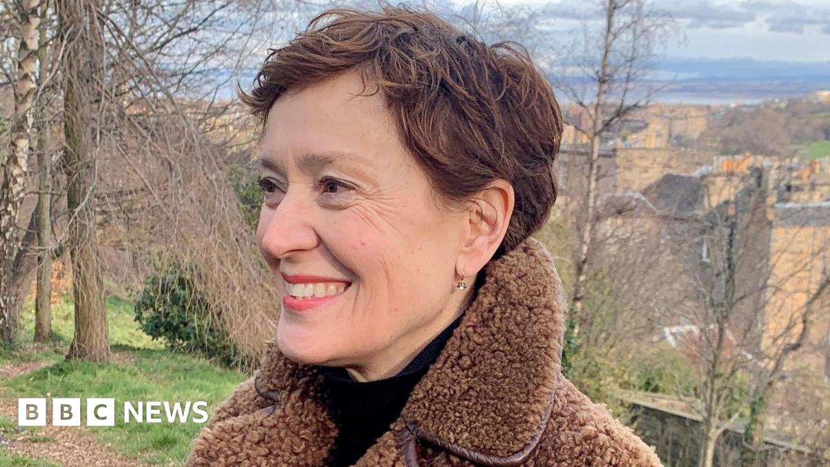 Carys Davies is smiling as she looks to the left of the camera. She has short brown hair and is wearing a black top with a brown fleece jacket. She is in the countryside, with trees behind her.