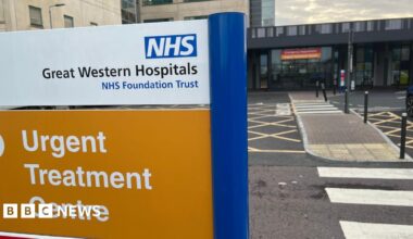 A sign outside a hospital building with the words "NHS Great Western Hospitals NHS Foundation Trust" and a second sign on an orange background below with the words "Urgent treatment centre". Hospital buildings and zebra crossings on the right.