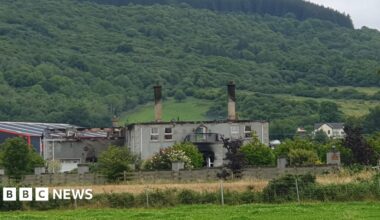 A house after a fire. No roof is left on the property. In the background, is hills and trees.