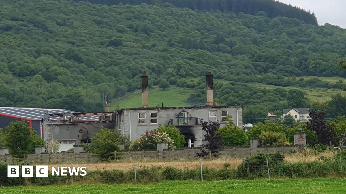 A house after a fire. No roof is left on the property. In the background, is hills and trees.