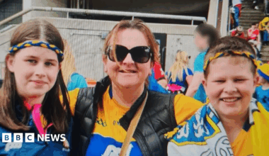 A woman stands in the centre. A girl is on one side and a boy on the other side. A lot of other people are behind them. The woman is wearing sunglasses and a GAA jersey. The boy and girl have blue and yellow bands on their heads. The boy has a flag draped over his neck. They're all smiling.