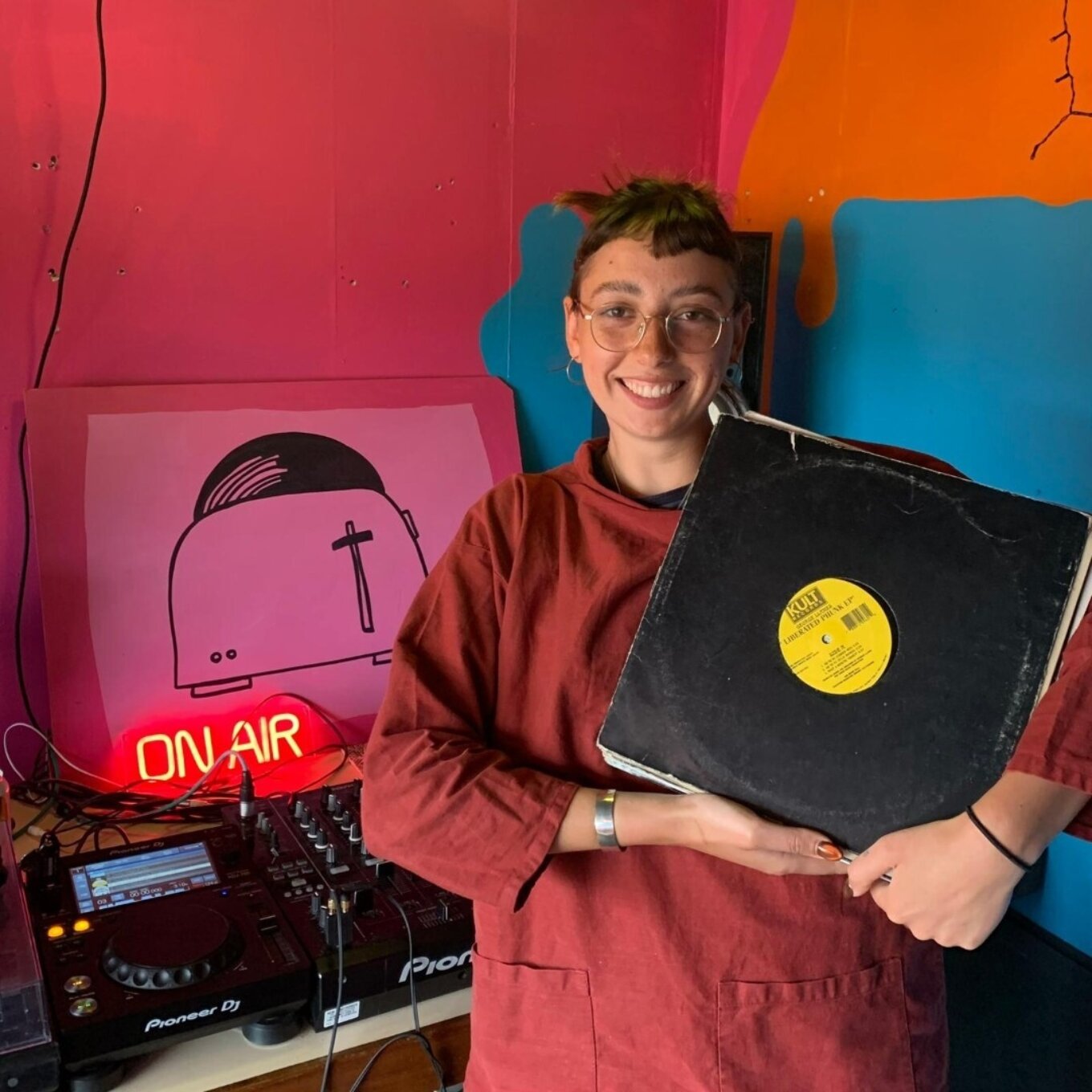 A photo of a smiling woman in a red overall holding an unidentified vinyl record. She's standing in a brightly painted room in front of a pair of DJ decks and a neon sign that says "ON AIR"
