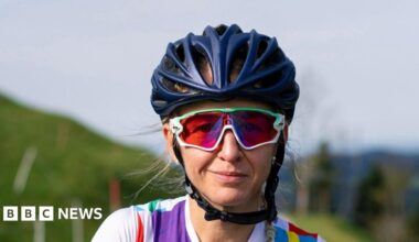 Emma Pooley is looking at the camera and is wearing a blue cycling helmet and glasses. She has long, blonde hair