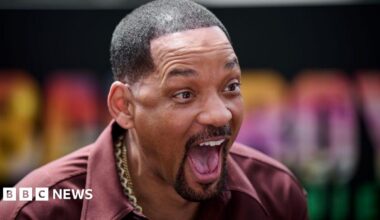 Will Smith is looking slightly to his right with an animated expression on his face: mouth wide open showing his tongue and upper teeth in a broad smile with his eyebrows high on his forehead. He has short cropped hair shaved into sharp edges, and is wearing an open necked burgundy shirt with broad lapels. He has several thick gold chains around his neck.