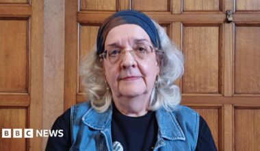 A headshot of an older woman. A polished wooden wall behind her suggests she is inside a town hall. building. She has a neutral expression and looks directly into the camera. She has grey shoulder length hair and wears glasses, hooped earrings, a navy bandana, black top and denim waistcoat.