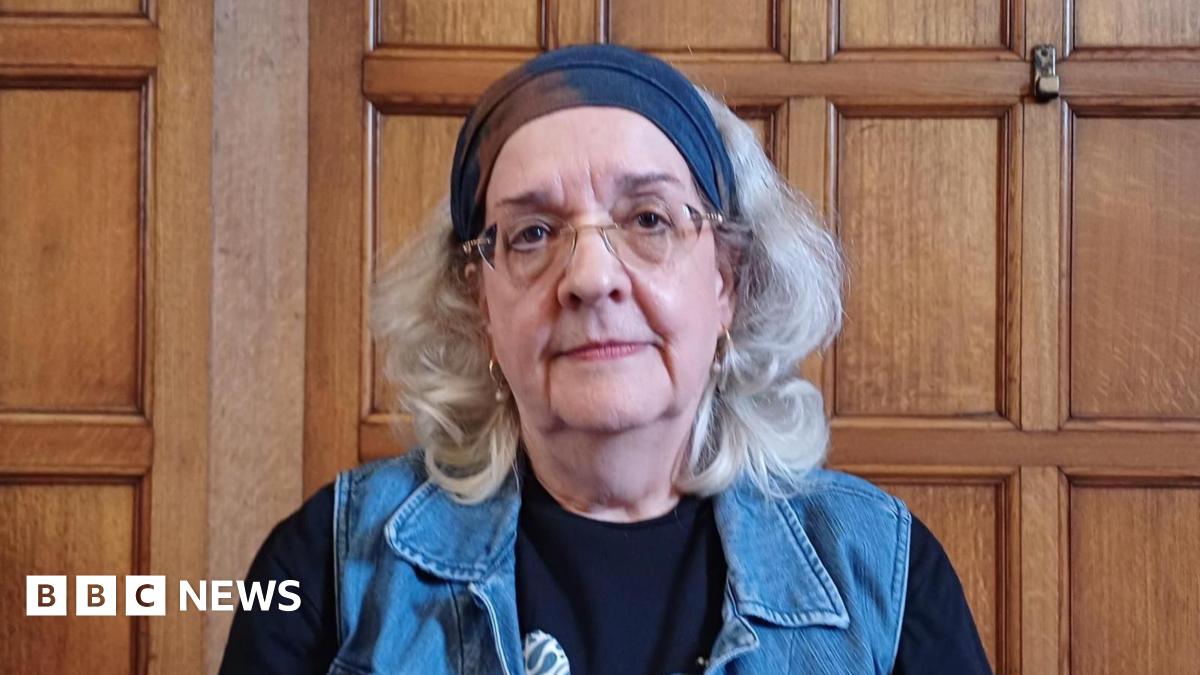 A headshot of an older woman. A polished wooden wall behind her suggests she is inside a town hall. building. She has a neutral expression and looks directly into the camera. She has grey shoulder length hair and wears glasses, hooped earrings, a navy bandana, black top and denim waistcoat.