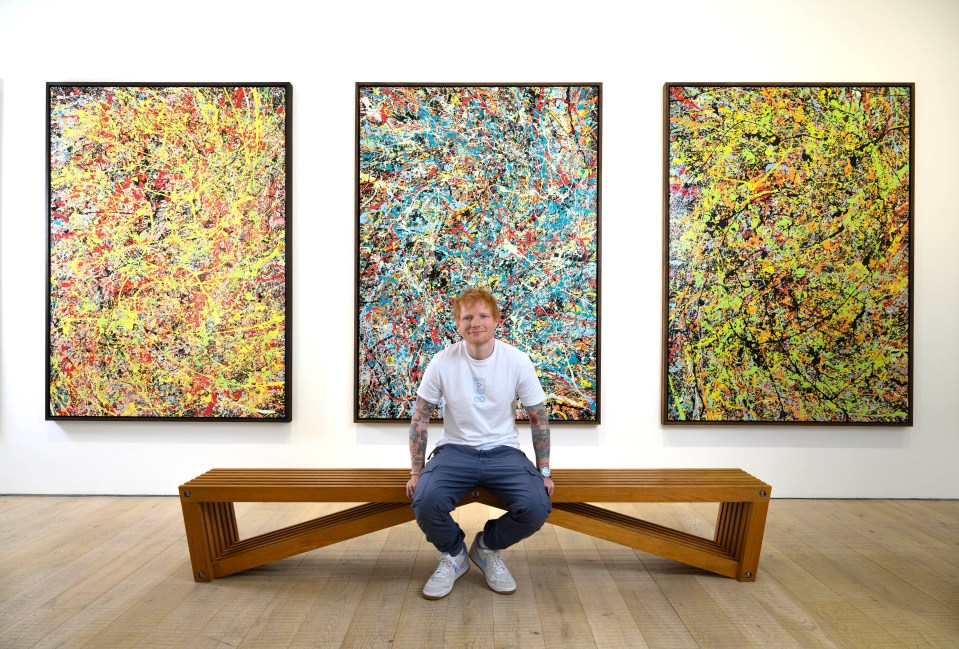 Ed Sheeran sitting on a bench in front of three of his paintings.