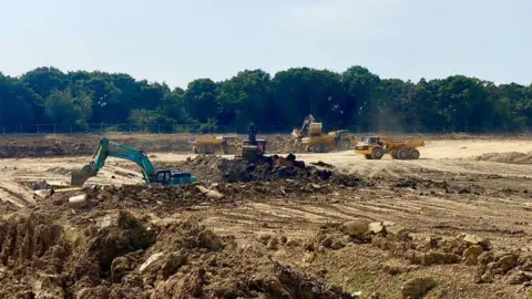John Fairhall/BBC Diggers churn up a large muddy plot of land on a sunny day.