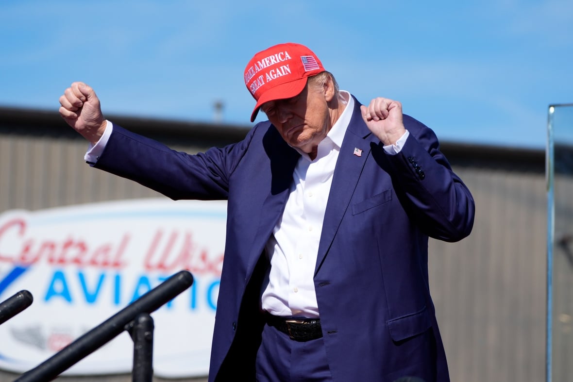 Donald Trump, wearing a a blue suit and a red ballcap saying 'Make America Great Again,' holds his arms in the air as he dances.   