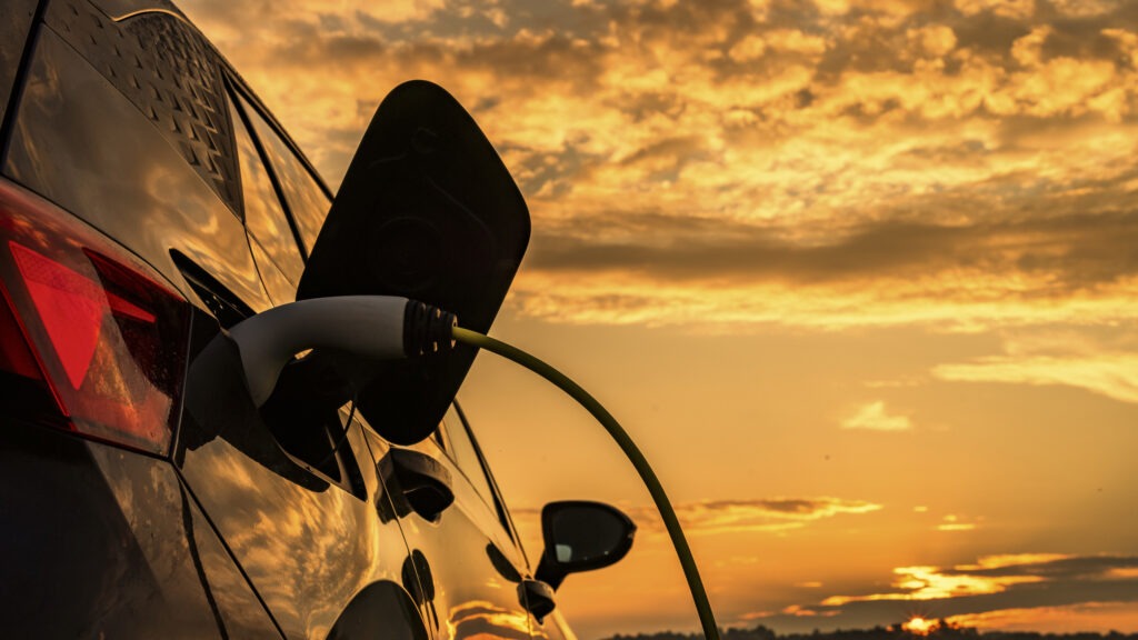 An electric car is plugged into a charging station as a glowing sunset sky reflects off its sleek metallic surface, symbolizing modern sustainability.