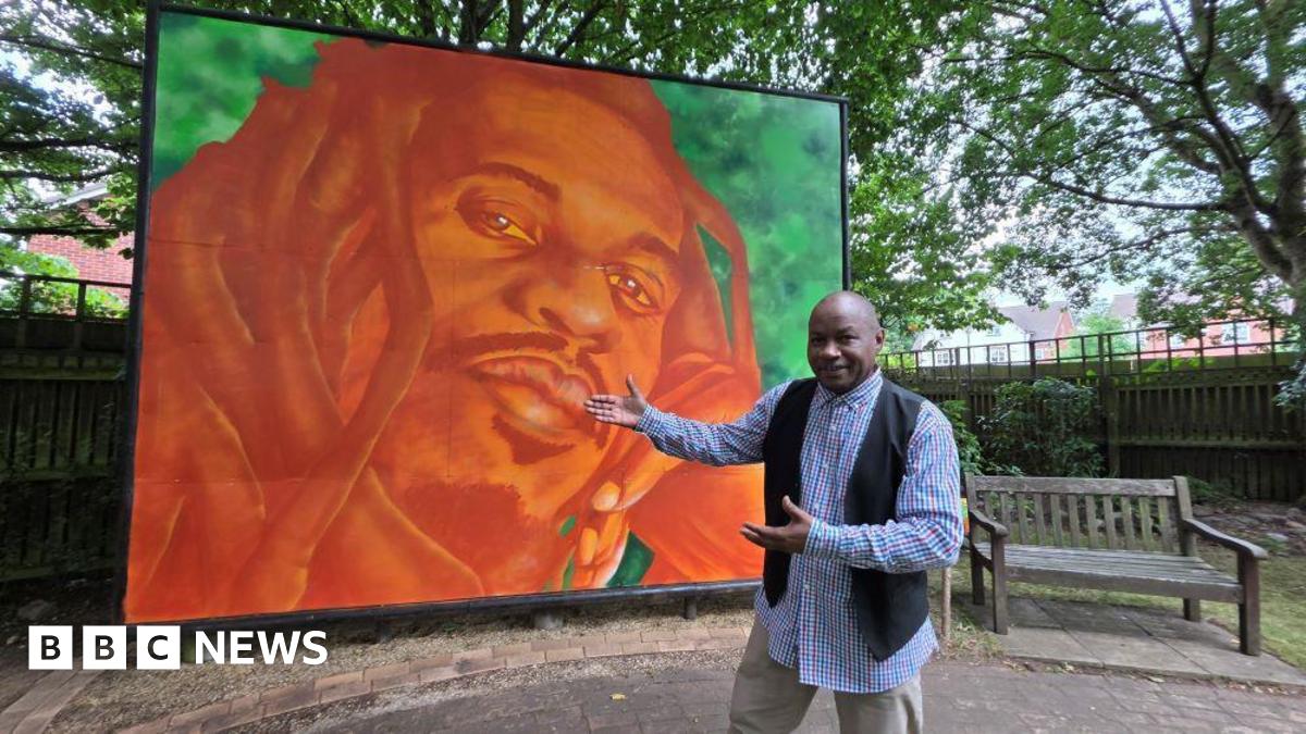 Tippa Naphtali wearing a red, blue and white checked shirt with a black waistcoat holds his arms out to present the new mural of his brother. The mural shows the face of Benjamin Zephaniah in reds and oranges in front of a green background.