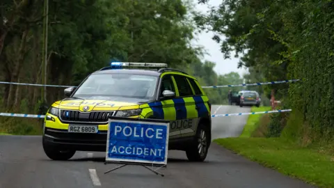 Pacemaker A blue sign in the middle of a road which read POLICE ACCIDENT - trees and grass verge on side of the road. Behind the sign is a police car which blue lights with a tape behind. 