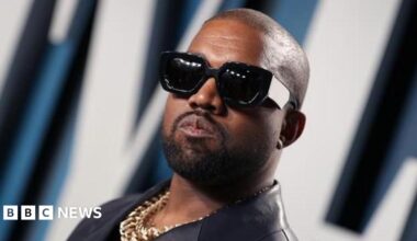Kanye West with sunglasses on posing for a picture. He is also wearing a gold chain.