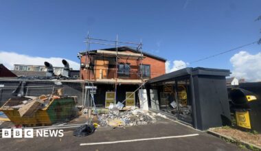 A restaurant destroyed by an arson attack. A small two storey-building is seen with rubble piled in front of it, black staining on the red bricks from the smoke and scaffolding up against the building. A skip is also outside the premises piled with rubbish.