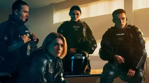BBC Series still of Blue Lights. Two men and two women in PSNI gear are gathered around a desk and wearing PSNI tactical riot gear. The woman in the centre nearest the camera has a blonde bob and a serious impression. The other woman has short dark hair and is standing behind  her. The men are on the left and right of the image and both have short brown hair.