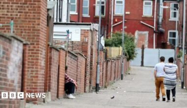Backstreets and alleys in the Eastwood area of Rotherham, with a man and a woman walking, and a man sitting. A bird and some litter is in the street with many red brick buildings.