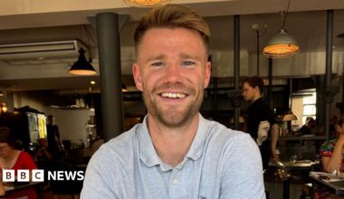 Hamish Douglas, a bearded man in his late 20s or early 30s smiling and wearing a grey polo shirt. He is sat inside a bar or restaurant, with industrial-style lighting behind him. Other customers can be seen at tables nearby.